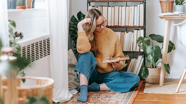 Woman sat on the floor in front of a bookshelf reading a book